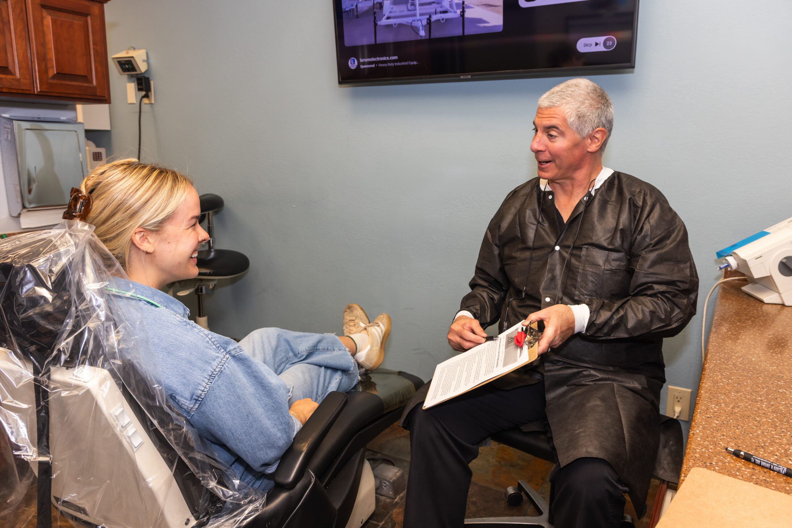 A woman in a dental chair smiles at a dentist who holds a clipboard